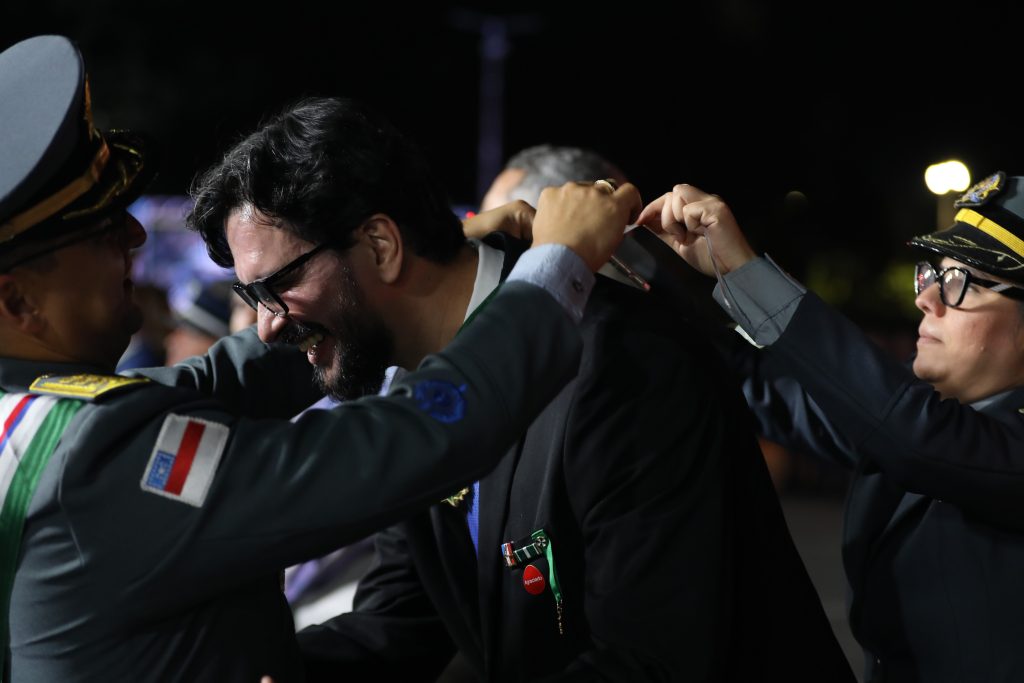Fotografia de plano médio tirada à noite durante solenidade oficial. No centro, o professor Denison Melo de Aguiar, de óculos e paletó escuro, sorri enquanto recebe uma medalha. À esquerda, um oficial da Polícia Militar do Amazonas (PMAM), fardado com quepe cinza e faixa transversal colorida, coloca a honraria em seu pescoço. À direita, uma oficial fardada de óculos auxilia na colocação da medalha. O ambiente está iluminado por refletores e o fundo está desfocado.