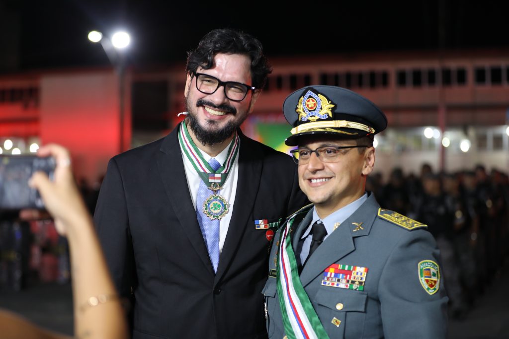 Fotografia de plano médio da solenidade oficial da PMAM. Em primeiro plano, à esquerda, as mãos de uma pessoa seguram um smartphone na horizontal, capturando uma foto da cena principal. Ao centro, o oficial da PMAM e o professor da UEA, Denison Melo de Aguiar, posam sorridentes lado a lado. O professor exibe a Medalha Ordem do Mérito no pescoço. O fundo noturno está iluminado e desfocado, destacando a interação entre os presentes e o registo do momento.