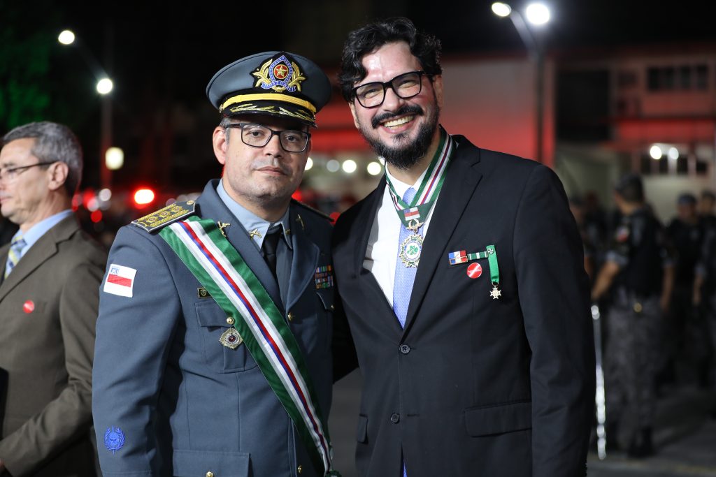 Fotografia de plano médio de dois homens sorridentes em pé, lado a lado, durante solenidade noturna. À esquerda, um oficial da Polícia Militar do Amazonas (PMAM) farda-se com uniforme cinza, quepe oficial e uma faixa transversal com as cores da bandeira do Amazonas. À direita, o professor da UEA, Denison Melo de Aguiar, veste paletó preto, gravata azul clara e exibe no pescoço a Medalha Ordem do Mérito – Grau Grã-Cruz, suspensa por uma fita listrada. O fundo mostra luzes da cidade e outras pessoas desfocadas, indicando um evento oficial ao ar livre.