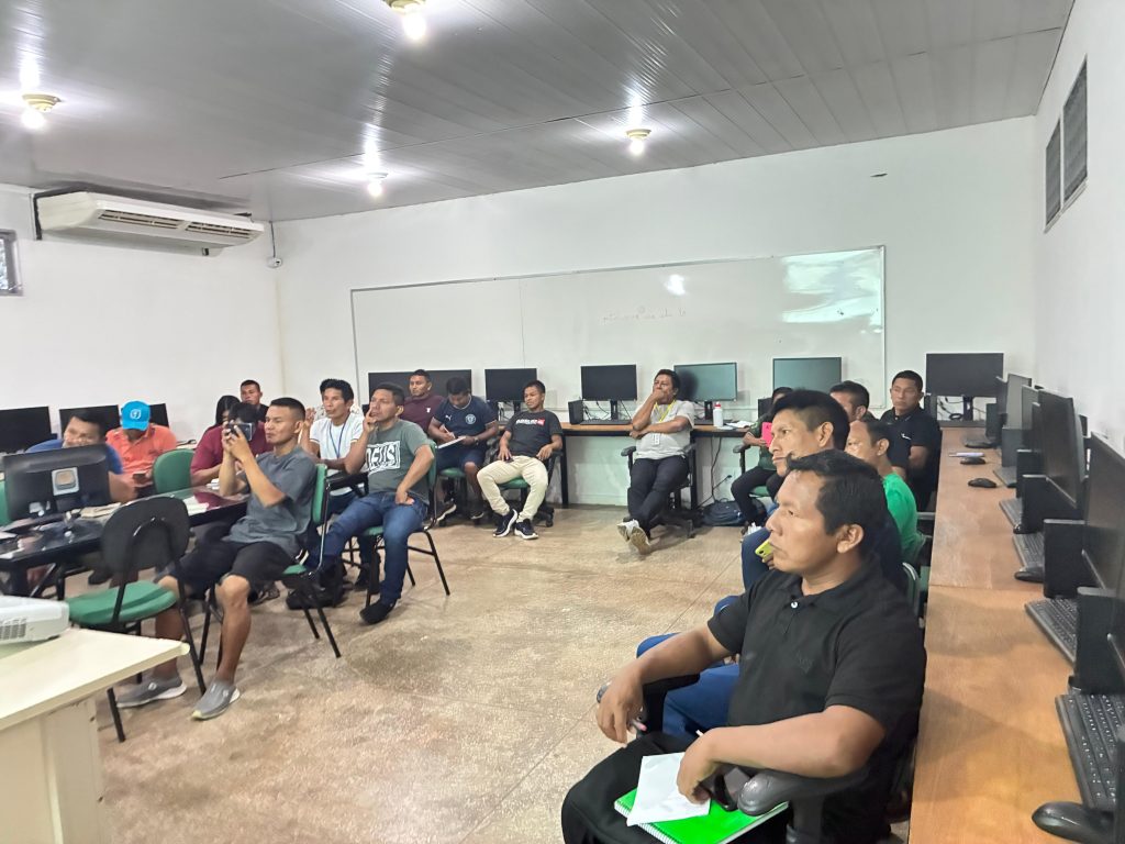 Vista ampla da sala de informática durante a capacitação. Diversos professores indígenas estão sentados em cadeiras verdes, organizados em torno de mesas com computadores. No centro, um dos participantes usa o celular para fotografar a apresentação. À direita, uma bancada de madeira exibe vários monitores alinhados. O ambiente possui paredes brancas, teto de PVC cinza e ar-condicionado ligado.