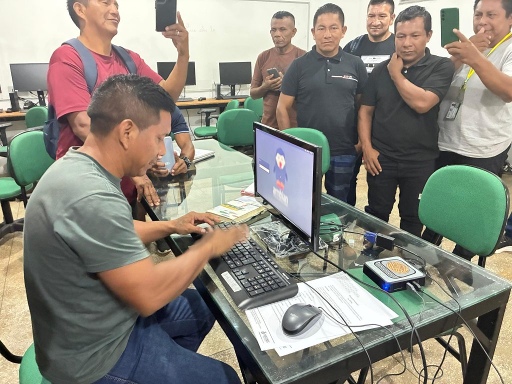 Um professor indígena de camiseta cinza está sentado à mesa de vidro, operando um computador com as mãos no teclado. À sua direita, sobre a mesa, está o dispositivo TV Box conectado ao monitor, que exibe uma interface gráfica colorida. Ao redor dele, um grupo de outros professores indígenas observa a tela com atenção; alguns ao fundo utilizam celulares para registrar a atividade. A cena ocorre em um laboratório de informática durante a oficina de capacitação da UEA para o uso educacional da tecnologia TV Box.