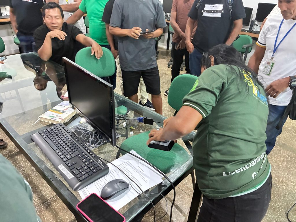 Vista superior em diagonal de uma mesa de vidro em um laboratório de informática. Uma mulher de cabelos pretos e camiseta verde está inclinada sobre a mesa, manuseando um pequeno dispositivo eletrônico preto conectado ao computador. Ao redor dela, um grupo de homens indígenas observa a ação. Sobre a mesa, veem-se um monitor, teclado preto, mouse cinza e documentos impressos. Outras pessoas aparecem em pé ao fundo, mantendo o foco na atividade técnica realizada na mesa.